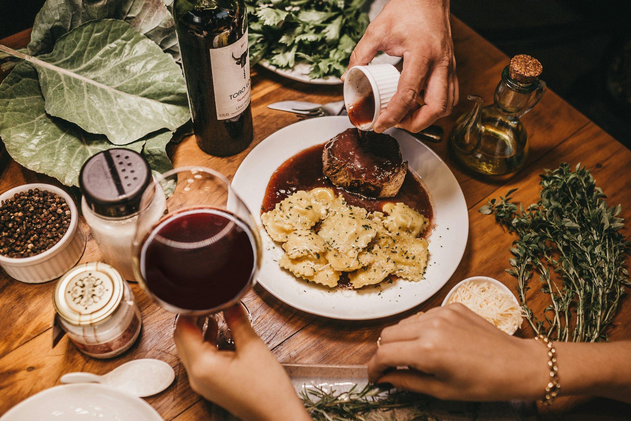 Hands serving steak and pasta at a rustic wooden table with a glass of red wine and fresh herbs