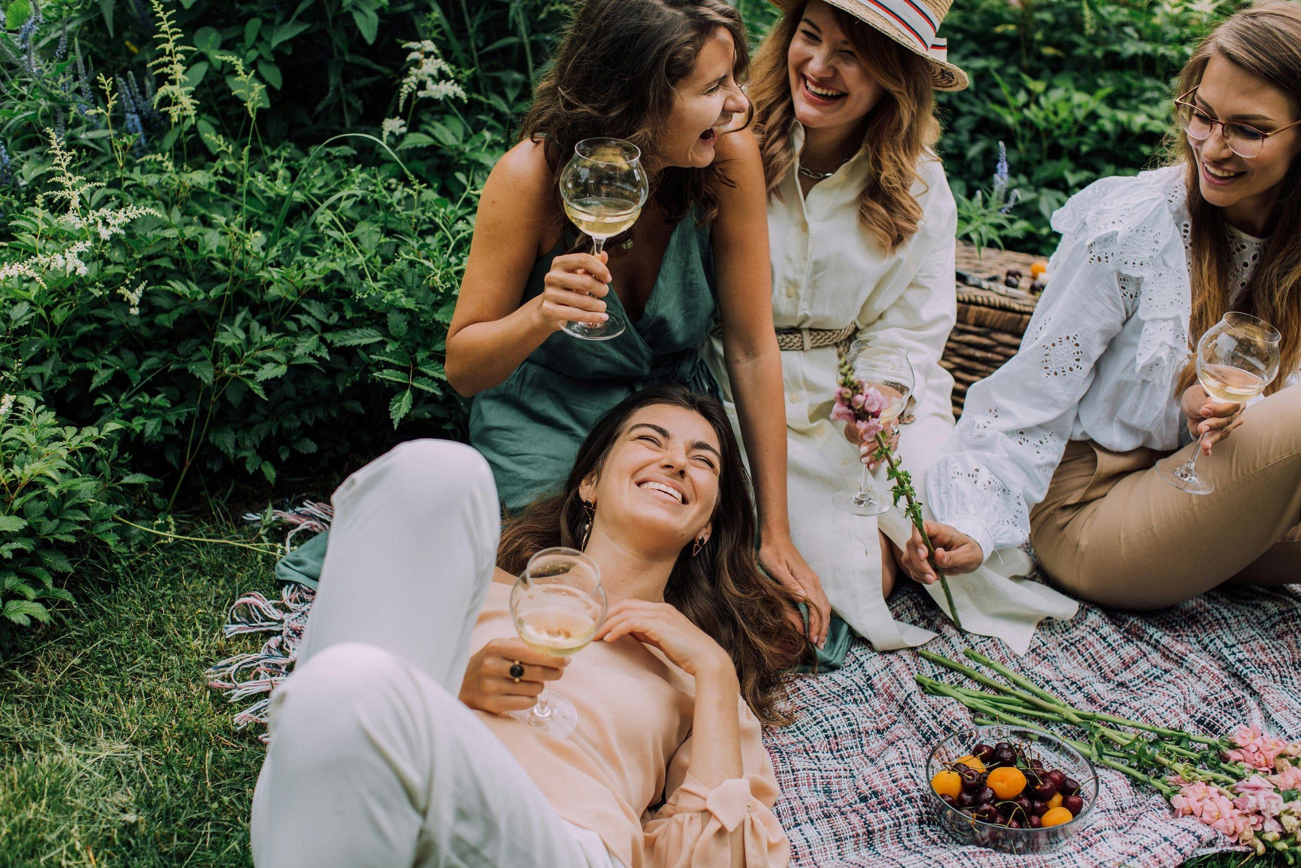 Four friends laughing together over glasses of white wine at a summer garden picnic