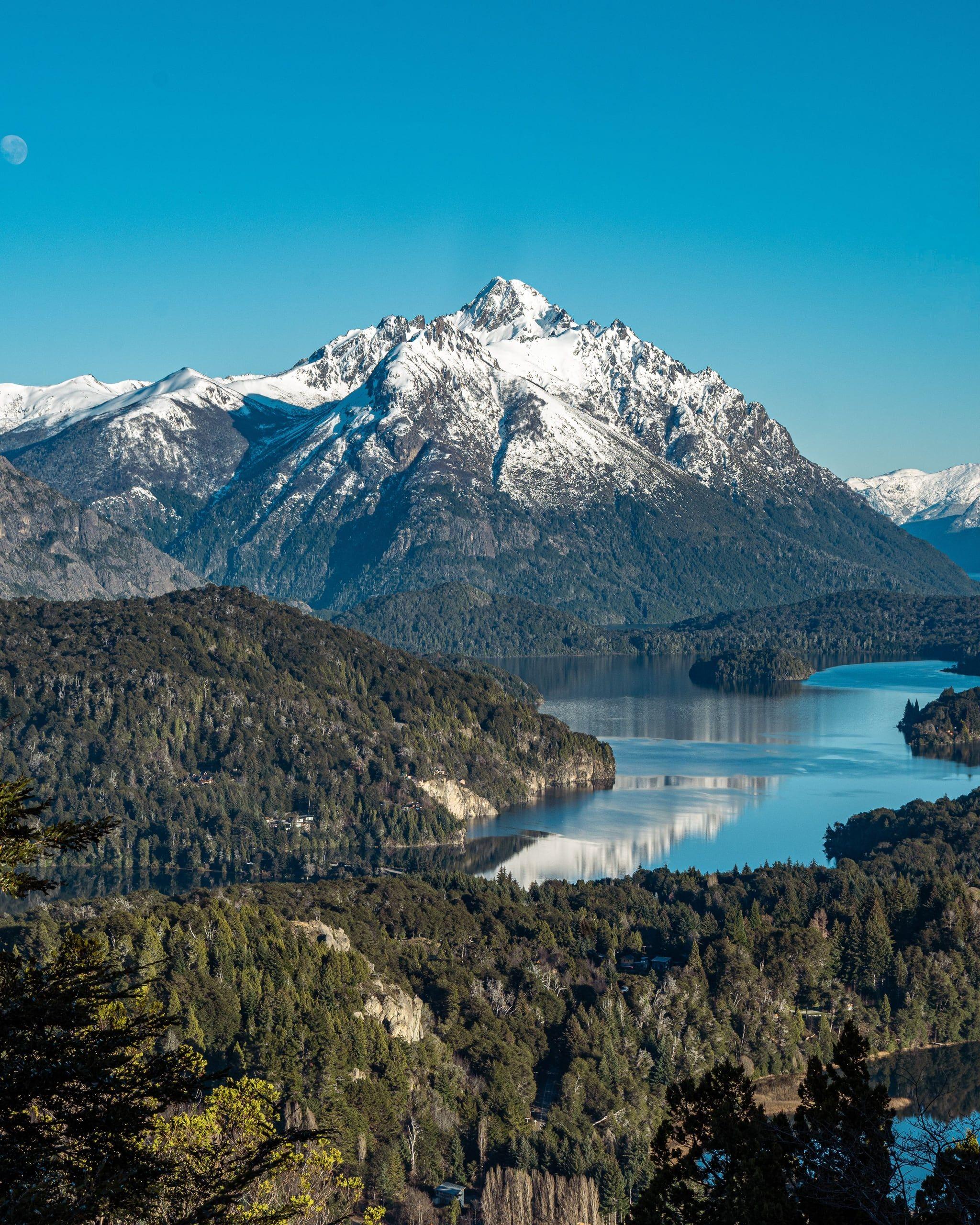 A snow-capped peak above a still lake and forested hills in Patagonia