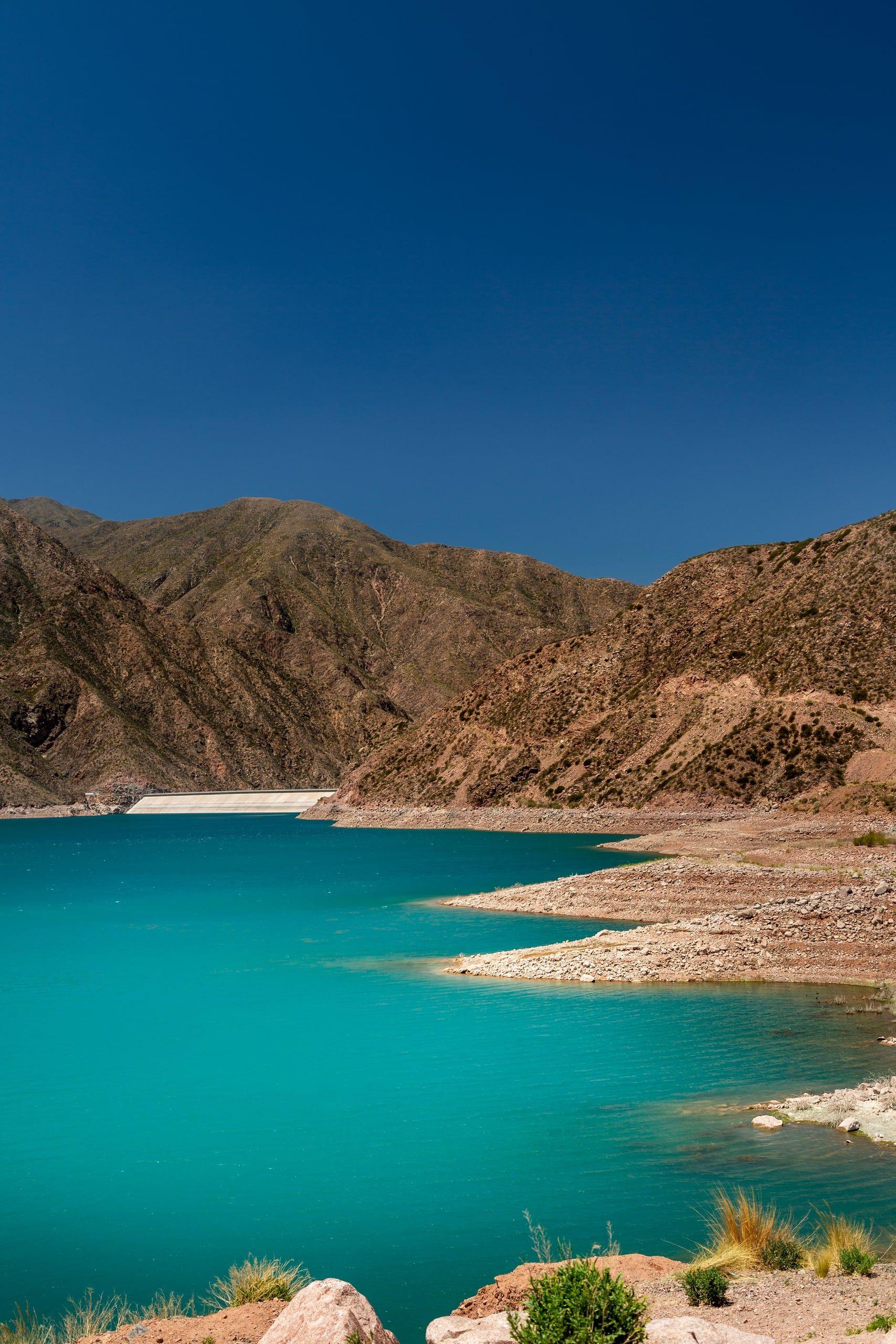 A turquoise reservoir held between the arid brown mountains of the Mendoza precordillera under a deep blue sky